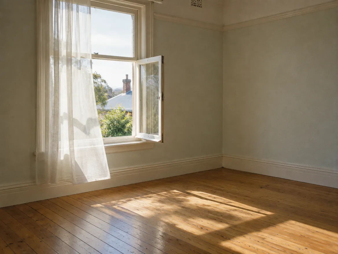 A quiet, empty room with timber floors and an open window letting in soft light.